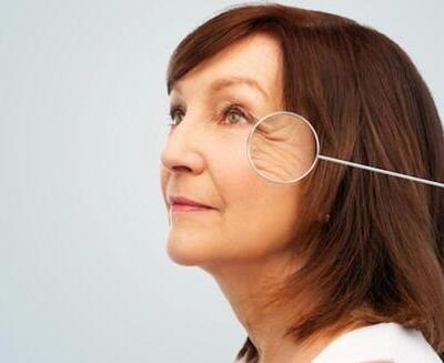 Mujer de mediana edad con pelo castaño mira hacia arriba; una lupa resalta las arrugas alrededor de su ojo derecho sobre un fondo claro liso.