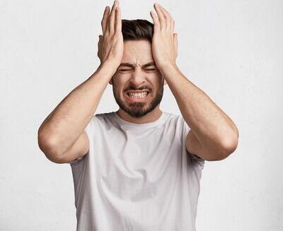Hombre con camiseta blanca de pie sobre un fondo liso, con los ojos cerrados, apretando los dientes y presionando ambas manos a los lados de la cabeza, expresando frustración o estrés.