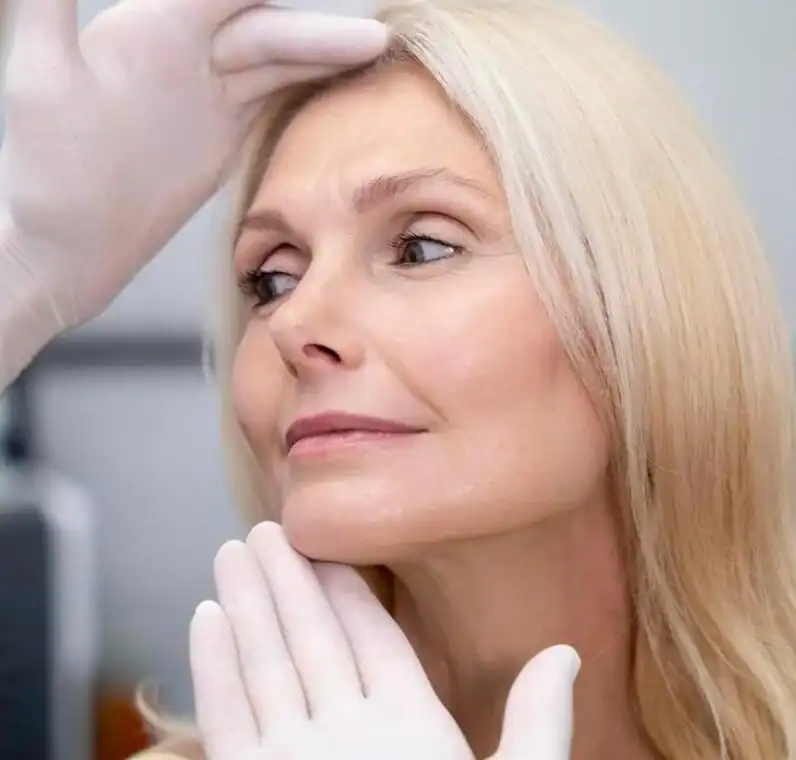 Se examina el proceso de medicina estética Tarragona, a una persona con pelo claro con las manos enguantadas, tocándole con una mano la frente y con la otra debajo de la barbilla, en un entorno clínico o médico.