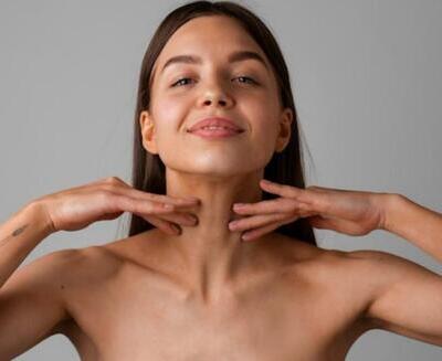 Una mujer de pelo castaño liso se toca el cuello con ambas manos, sonriendo ligeramente, sobre un fondo gris liso.