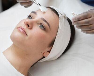 Una persona tumbada con una cinta en la cabeza recibe un tratamiento facial mientras una mano enguantada le aplica un producto en la frente con un cepillo.