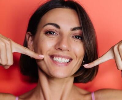 Mujer de pelo castaño corto sonríe y se señala los dientes con ambos dedos índices; fondo naranja.