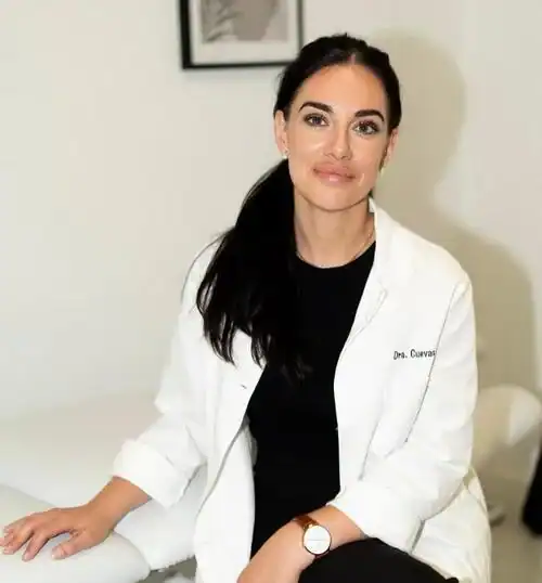 Dra. Lidia Cuevas de pelo largo y oscuro, con bata blanca de laboratorio, se sienta en una silla de un despacho luminoso y minimalista.